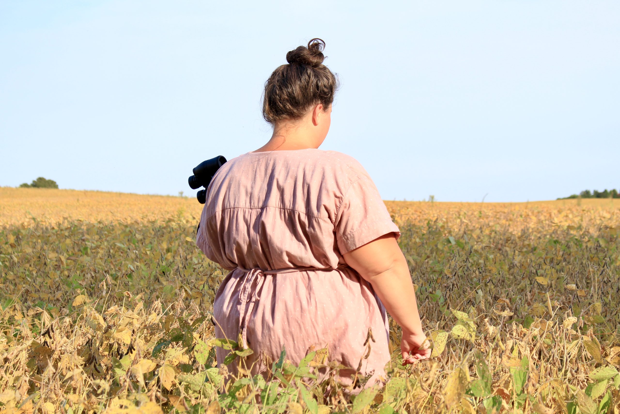 woman with a larger body standing in a field wearing a pinkish custom made tunic with waist tie and holding a pair of binoculars.